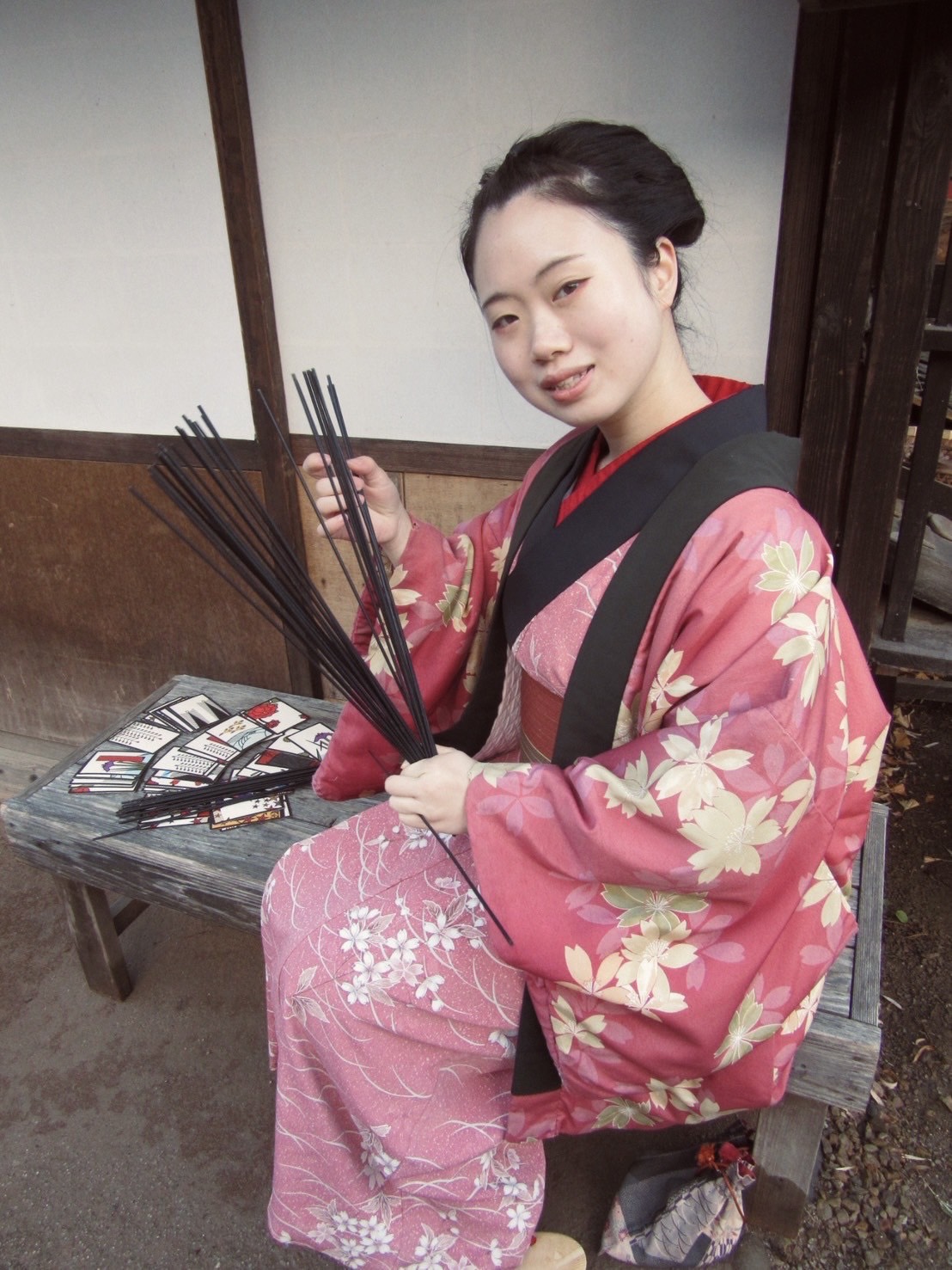 桜柄の着物で縁台に座り、筮竹を持って微笑む花緒よみ