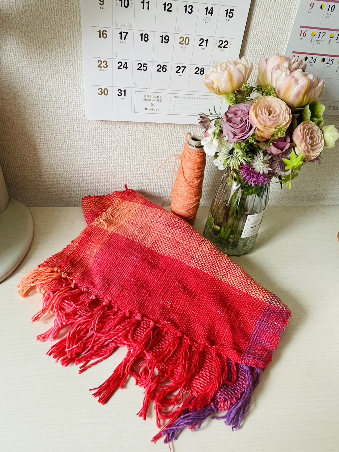 机の上に置かれた真紅から朱へ移る手織りストール。橙の糸巻きと花が添えられている