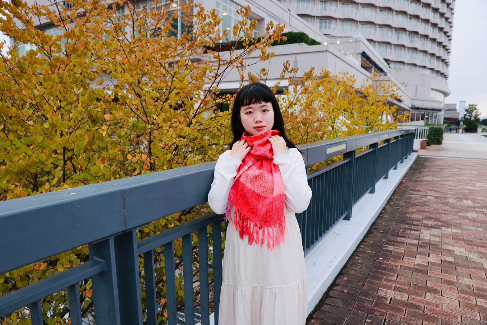 黄葉の並木を背に、真紅の手織りストールを手に取っている花緒よみ